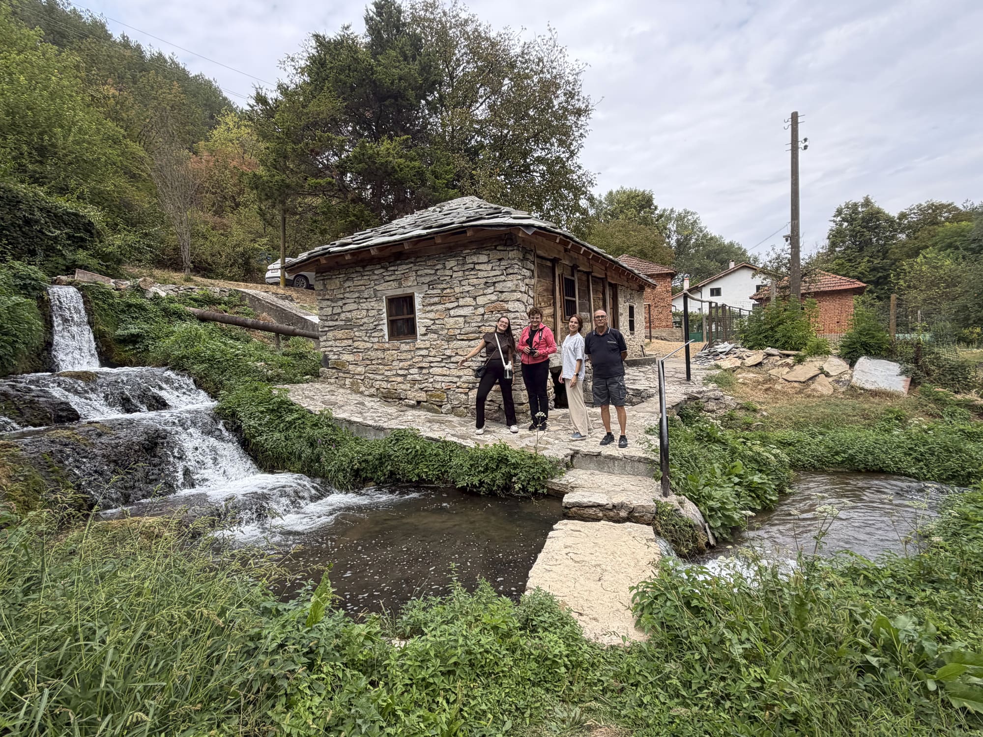 Банковската воденица, каменна сграда с покрив от каменни плочи край реката, с водата, падаща по прага до нея, и четирима души пред вратата